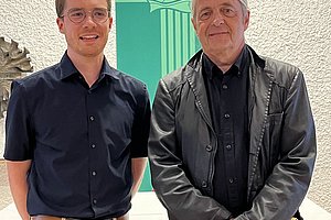 Zwei Personen stehen nebeneinander, beide tragen dunkle Hemden. Der Mann links hat eine hellbeige Hose an, der Mann rechts trägt eine schwarze Jacke. Im Hintergrund hängt ein grünes Banner mit einem dekorativen Muster.