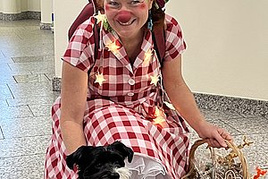 Eine sitzende Person in kariertem Kleid und roter Baskenmütze hält einen Korb mit Lebkuchenherzen, neben ihr sitzt ein kleiner Hund mit blauem Halstuch