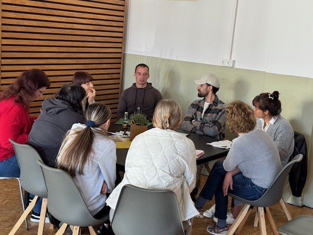 Acht Personen sitzen im Kreis auf Stühlen und führen eine Gruppendiskussion in einem Raum mit Holzpaneelen an der Wand.