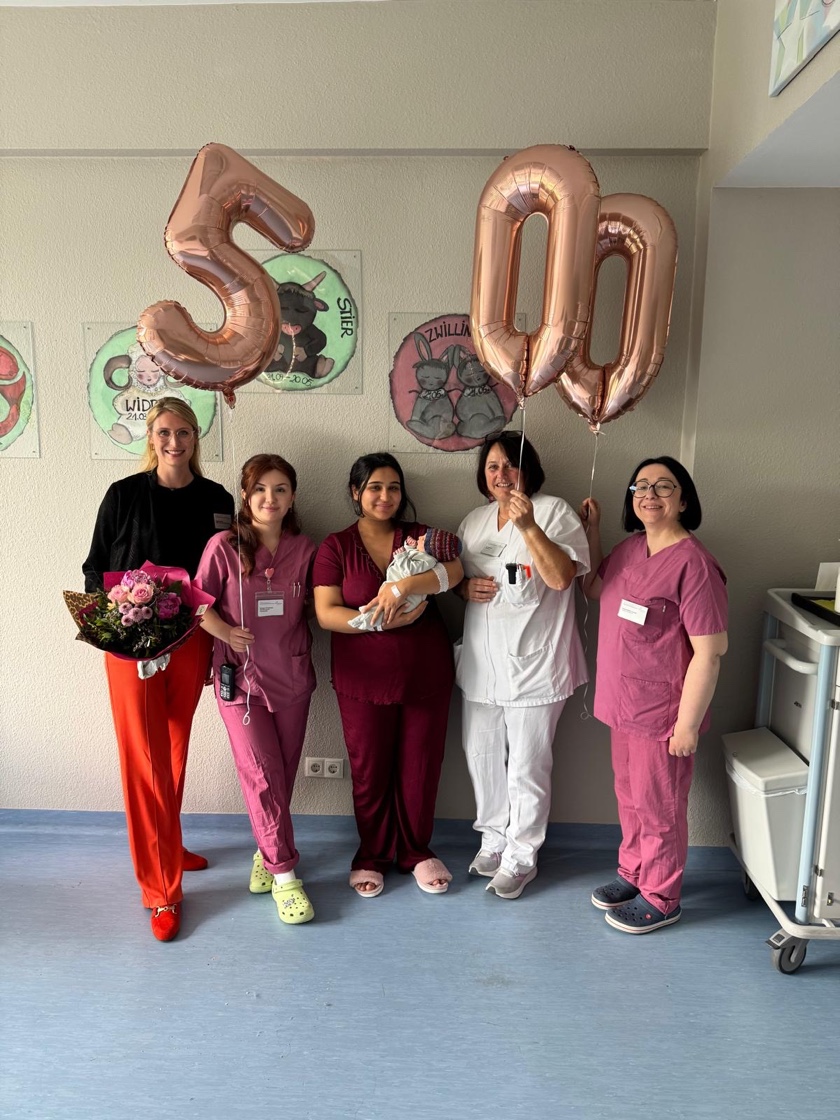 Fünf Personen in medizinischer Kleidung stehen in einem Raum, zwei halten große roségoldene Luftballons in Form der Zahlen 500, eine hält einen Blumenstrauß, eine weitere einen Babystrampler