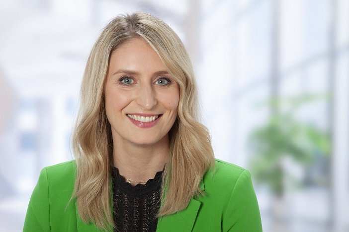 Eugenia Kompe mit langen blonden Haaren, trägt grünen Blazer und schwarze Bluse, vor hellem Bürohintergrund